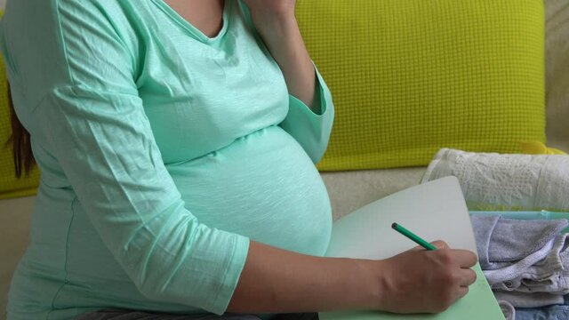 Cute Pregnant Woman writing packing list for maternity hospital With Notebook Prepares bags. Young Ledy In Pregnancy Have Fun Spend Time On Sofa At Home. Motherhood, Medicine Health and Care Concept