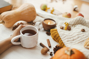 Obraz na płótnie Canvas Hello autumn, cozy slow living. Hand holding warm cup of tea on background of autumn leaves, pumpkin, cozy sweaters, burning candle on white wood. Happy Thanksgiving