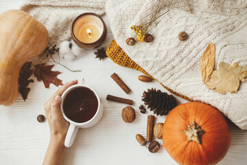 Obraz na płótnie Canvas Hello autumn, cozy slow living. Hand holding warm cup of tea on background of autumn leaves, pumpkin, cozy sweaters, burning candle on white wood. Happy Thanksgiving. Flat lay