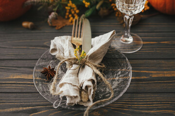 Thanksgiving dinner rustic table setting. Modern plate with vintage cutlery, linen napkin, herb and glass on wooden table with pumpkins and autumn flowers arrangement. Eco friendly catering