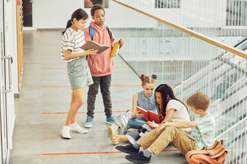Full length portrait of diverse group of children reading books while waiting in hall of modern school, copy space