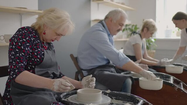 A female potter's wheel sculpting teacher explains how to work and teaches an elderly woman to work with clay and make mugs and jugs. Master class for pensioners. Pottery courses
