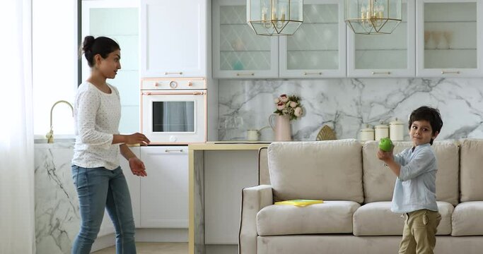 Indian woman play with 4s son in modern apartment throwing each other green apple enjoy playtime spend free time at cozy home. Healthy eating and lifestyle, lively games with children indoor concept