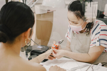 Asian manicure therapist wearing face mask filing customer nails in nail salon, with protective screen, social distancing covid-19 pandemic procedure, reopenning small business