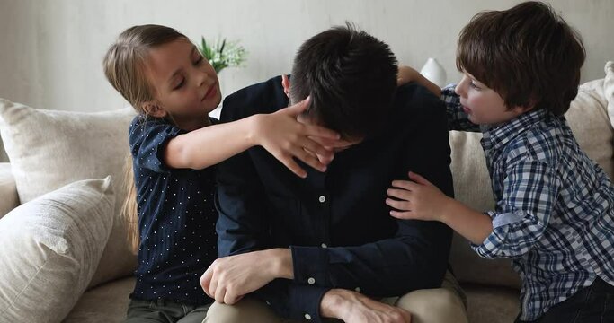 Stressed unhappy young single father irritated by naughty small preschool children, needs rest sitting on sofa. Adorable little brother and sister cuddling, communicating with depressed millennial dad