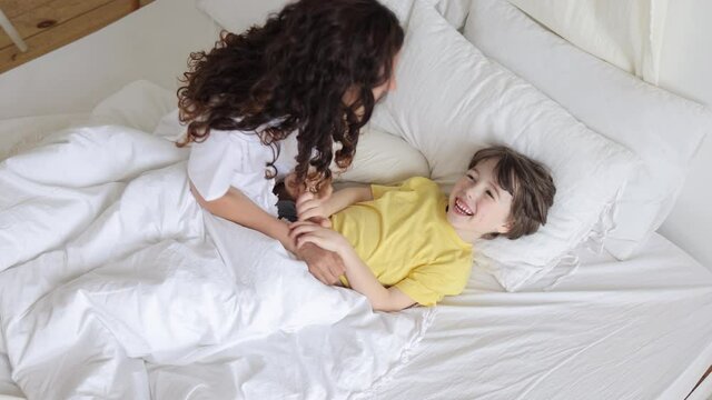 Happy mom play with little child tickling laughing boy lying in bed. Caring cheerful mother waking up kid on morning weekend with funny active game. Mum and son loving spend time together in bedroom