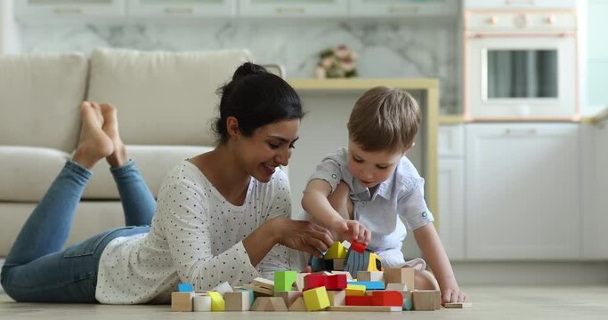 Indian female nanny play wooden colour cubes with 5s Caucasian boy, people enjoy playtime on warm floor in cozy living room. Children development, toys shop, weekend leisure with kids at home concept