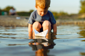Small caucasian boy blonde hair three years old sitting alone on the coast of the river or lake in sunny summer or autumn evening playing with paper boat childhood tranquility concept copy space