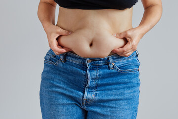 overweight woman in jeans with a bare dangling belly. Studio photo on gray background