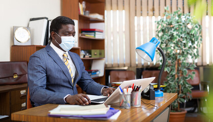 Focused african american businessman in a protective mask, working for a company during a pandemic, works at a computer..in the office