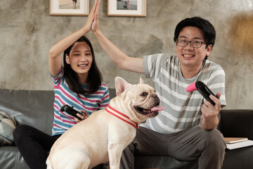 Asian couple is playing video games console, excitedly and having fun on sofa at home, control joystick to competition, pet dog (French Bulldog) nearby. It's happiness in family lifestyle.