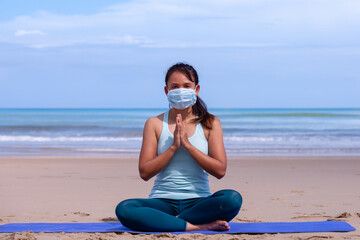 people, fitness, sport and healthy lifestyle concept - young asian girl making maditation in medical mask on tropical beach