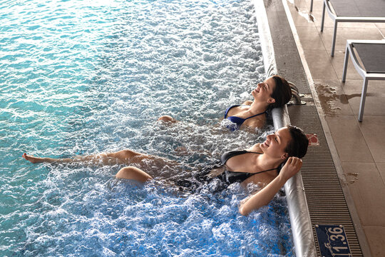 Two happy attractive brunette female friends in bikini relax and enjoy in air bubbly water in hotel swimming pool. Above view.