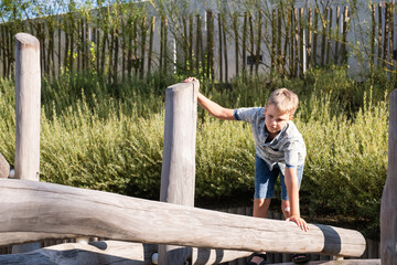 Blond boy walks on wooden beam in a playground in a public park.