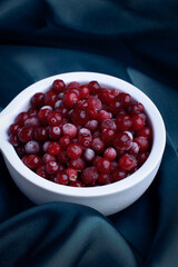 Frozen cranberry in white bowl on blue silky background. Selective focus. Close up of frozen cranberries.