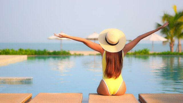 Backside view of sexy woman sitting on the side of swimming pool at an exotic hotel in Bali in yellow monokini and hat lifting her hands up, she up her arms wide open, blurred palm and beach umbrellas