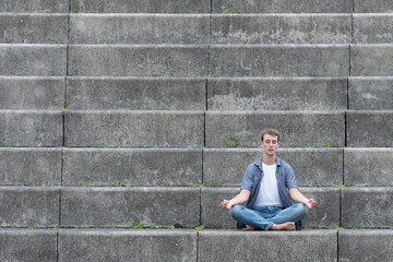 Fototapeta na wymiar Casual dressed young man sitting on stairs and meditating. Mental health, mindfulness and yoga concept with copy space.