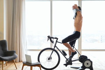 Fototapeta na wymiar Athletic young man taking off shirt while cycling indoors at home