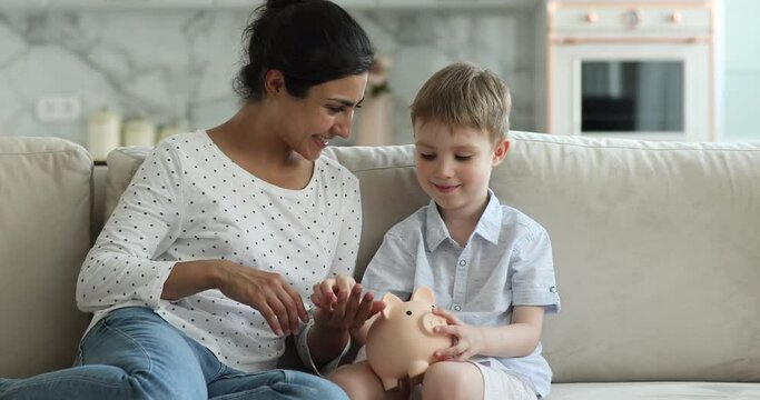 Little Caucasian boy sit on sofa at home takes coins from Indian mother and drops it into piggy bank, babysitter teach child to be thrifty. Make preparation for future of kid, deposit, savings concept