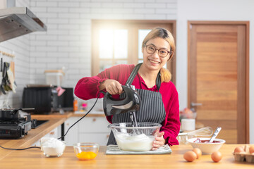 Asian girl using the blender, blend dough and other ingredients for cooking the chocolate cake with happy feeling in the kitchen room.