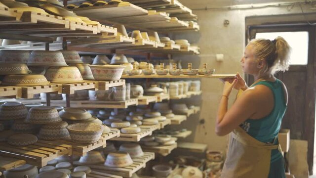 Female Pottery artist aranging clay dishware on wooden rack shelves