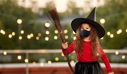 halloween, holiday and pandemic concept - girl in black protective mask and costume of witch with broom over garland lights at roof top party background