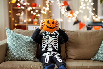 halloween, holiday and childhood concept - smiling little boy in costume of skeleton with jack-o-lantern sitting on sofa at home