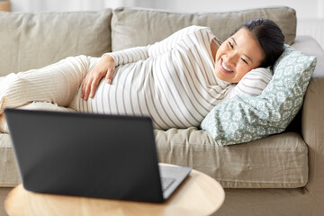 pregnancy, rest, people and expectation concept - happy smiling pregnant asian woman with laptop computer sitting on sofa at home