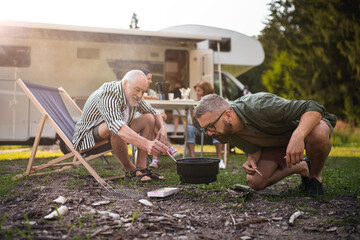 Mature man with senior father preparing barbecue at campsite outdoors, caravan family holiday trip.