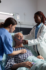 African doctor and medical assistant helping senior man breath using oxygen mask, in hospital laying in bed. Sick patient cant breath because of lung infection. Nurse listening heart with stethoscope.