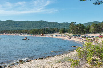Fototapeta na wymiar Scenic public beach with swimming and relaxing vacationers and natural coniferous green forest on hills, summer landscape of calm sunny bay