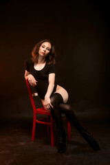 Fototapeta na wymiar A slender woman in a black bodysuit and stockings posing on a black background on a red chair.