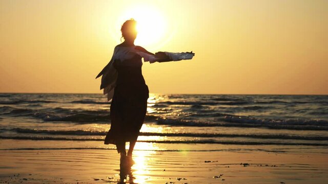 Silhouette of young girl with angel wings dancing at sunset. Silhouette of female artist is dancing on the sandy beach against orange sunset, slow motion