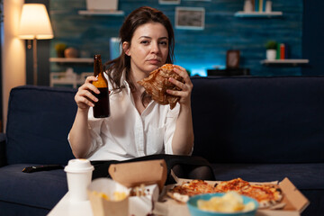 Portrait of caucasian woman looking into camera relaxing on sofa in living room enjoying takeaway food home delivery. Woman eating tasty delicious burger. Fastfood lunch meal order