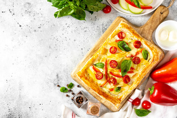 Puff pastry pie with mixed vegetables and mozzarella cheese on a serving board on a culinary light table top view. Delicious hearty pastries with tomatoes, zucchini, sweet pepper, spices and basil