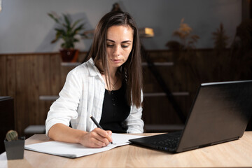 An adult college or university student learns remotely using a laptop and writing in a notebook. Online education or work at home. Caucasian girl writes homework in a notebook and studies