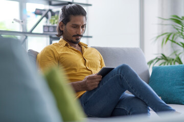 Fototapeta premium Indian man sitting on the couch a home and using a digital tablet