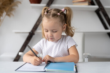 Naklejka premium Elementary school student preschooler studying at home does homework handwritten in a notebook or notepad sits at a desk. The preschooler learns online remotely. Distance education