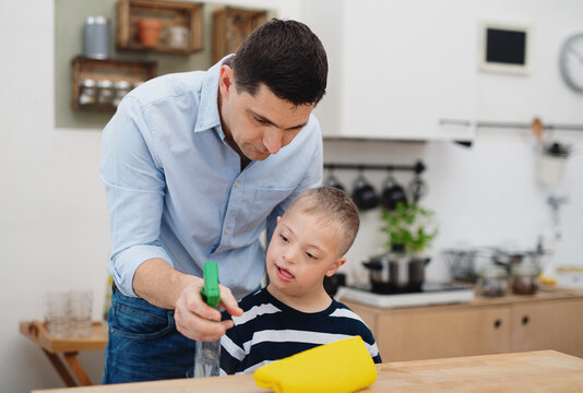 Father With Happy Down Syndrome Son Indoors In Kitchen, Cleaning Table.