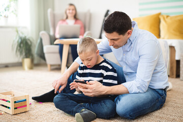 Father with happy down syndrome son indoors at home, using smartphone.