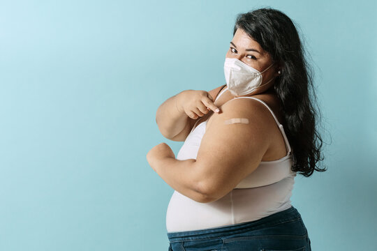 Oversized latinx woman using face mask pointing with her finger an bandage vaccine