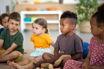 Group of small nursery school children sitting on floor indoors in classroom, listening to teacher.