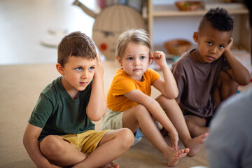 Group of small nursery school children sitting on floor indoors in classroom, listening to teacher.