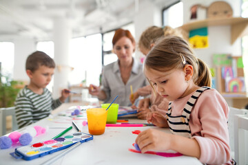 Group of small nursery school children with teacher indoors in classroom, painting. - obrazy, fototapety, plakaty