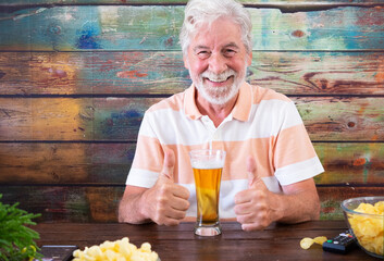 Happy senior white-haired man sitting at wooden table with beer and chips makes positive sign with...