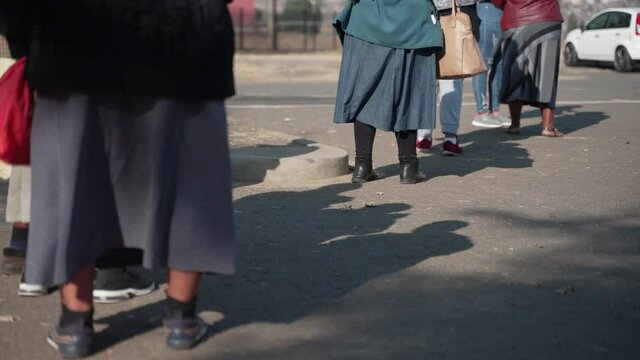Elderly African women stand in a long queue waiting outdoors, their skirts blow in the chilled breeze as the early morning sun casts their shadows on the asphalt ground.