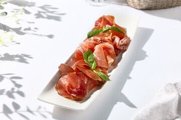 Italian antipasti on white table. Meat starter - sliced meat with basil on white dish. Sunshine and shadow on white table. Traditional italian first course include prosciutto, bresaola and parma ham.
