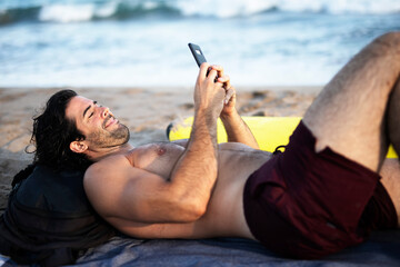 Handsome man resting on the beach, using the phone. Surfer taking a break on the beach..