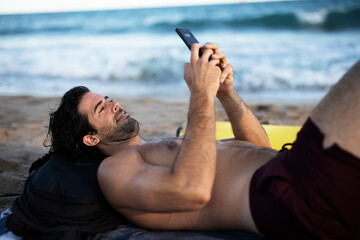Handsome man resting on the beach, using the phone. Surfer taking a break on the beach..