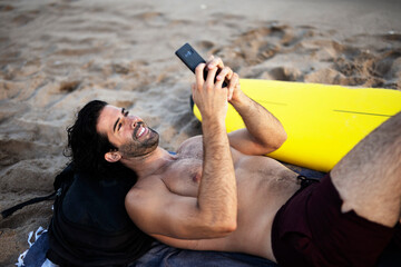 Handsome man resting on the beach, using the phone. Surfer taking a break on the beach..